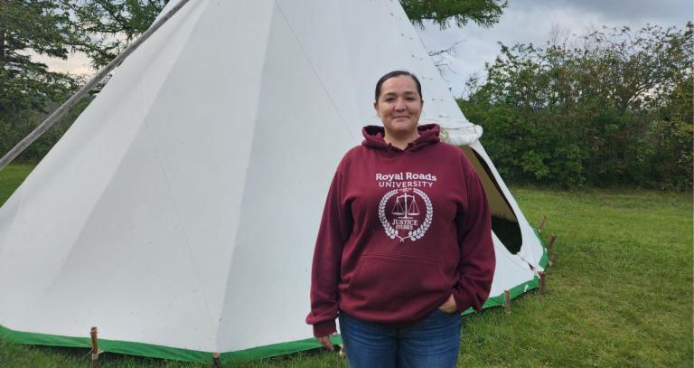 Dominique Bradford stands in front of a teepee on a grassy field. She wears a burgundy "Justice Studies" hoodie.