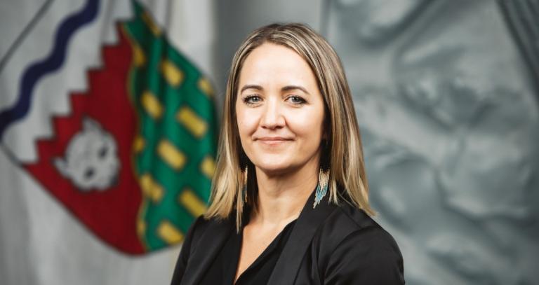 Kim Wickens smiles at camera. She has shoulder length blonde hair and is wearing a black shirt. A flag of the Northwest Territories is in the background.