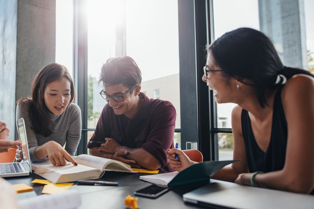 A table of three or four undergraduate students sit next to each other and look excitedly at a university textbook.