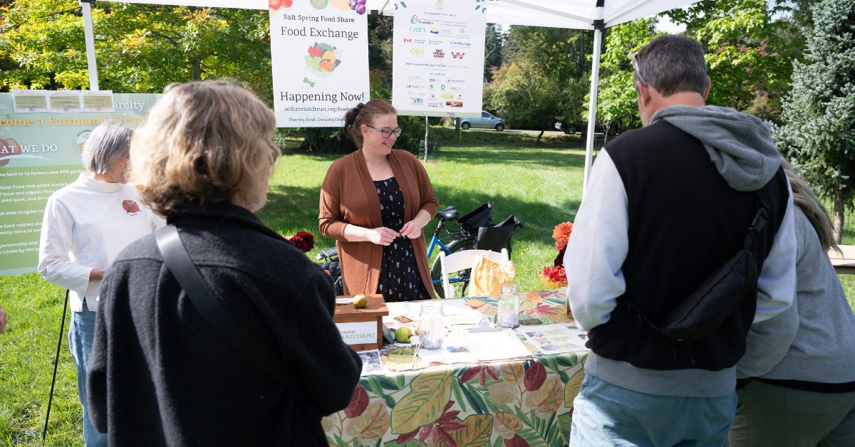 A group of people is gathered around an information table in an apple orchard. A woman is speaking to them. A sign saying Salt Spring Island Food Exchange is behind her.