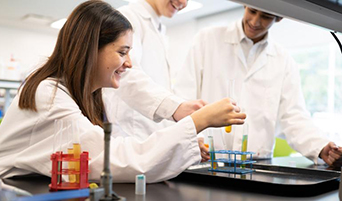 Three students wearing lab coats work together on an experiment, holding a test tube.