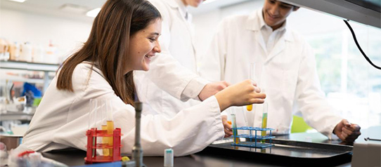 Three students hold up a test tube in a science lab wearing lab coats.