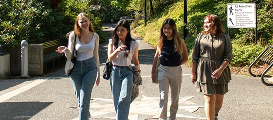 A group of four students walk through a paved path, surrounded by foliage at Royal Roads University