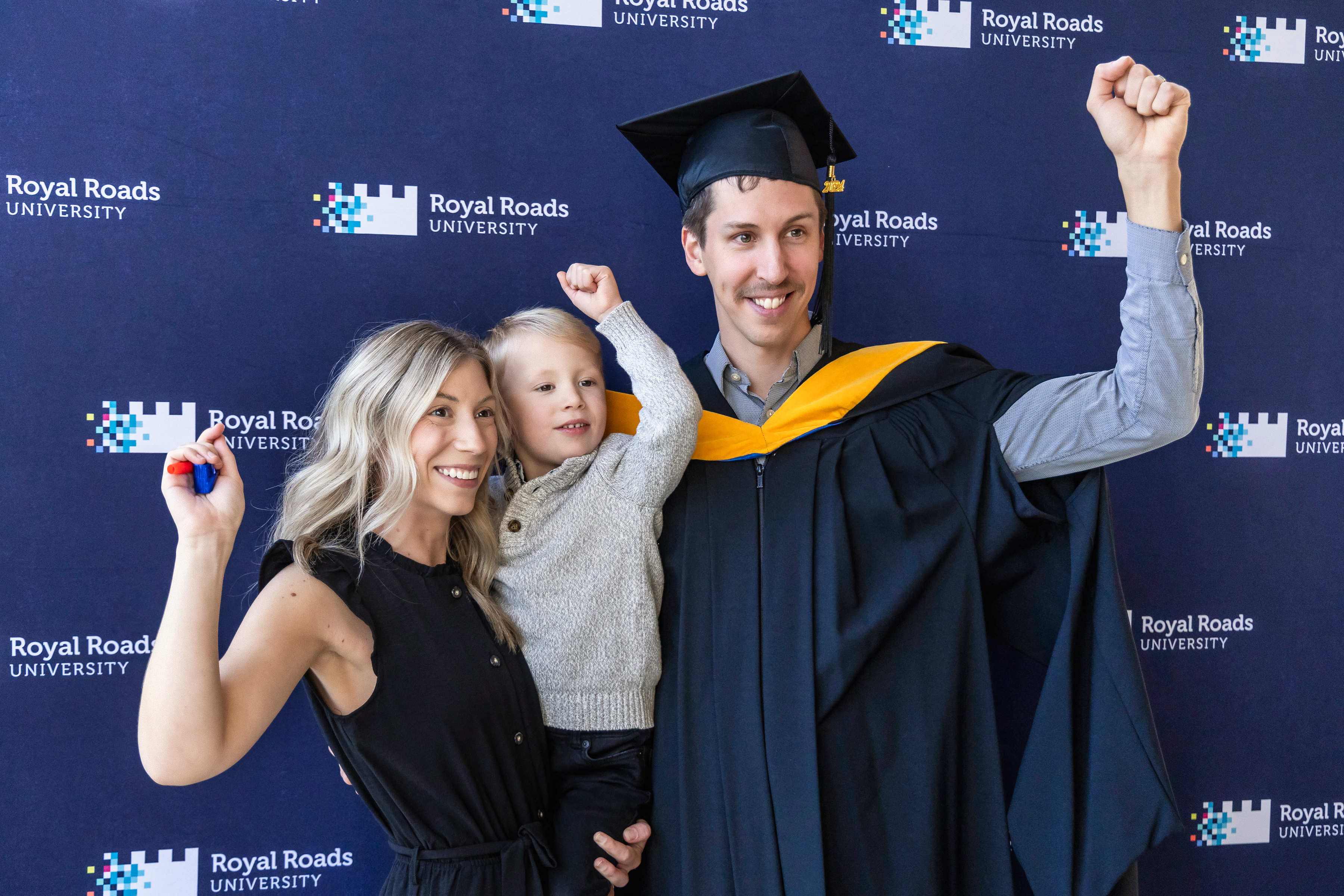 A young graduate with his family all raising their arms in joy against a branded RRU backdrop.