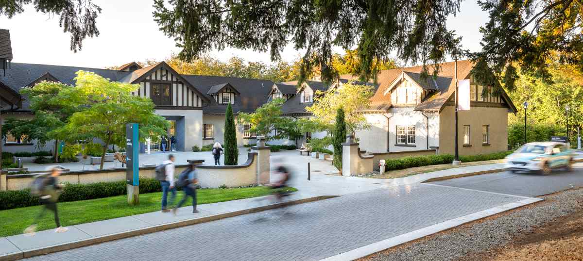 Students and cars pass by a building on a cobblestone road at Royal Roads University.