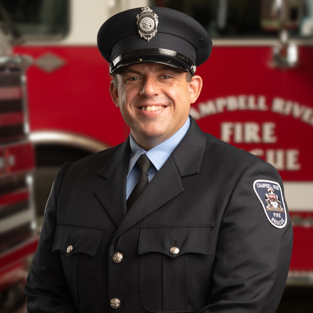 Jesse Challoner in dress uniform is smiling and standing in front of a Campbell River Fire Rescue Truck.