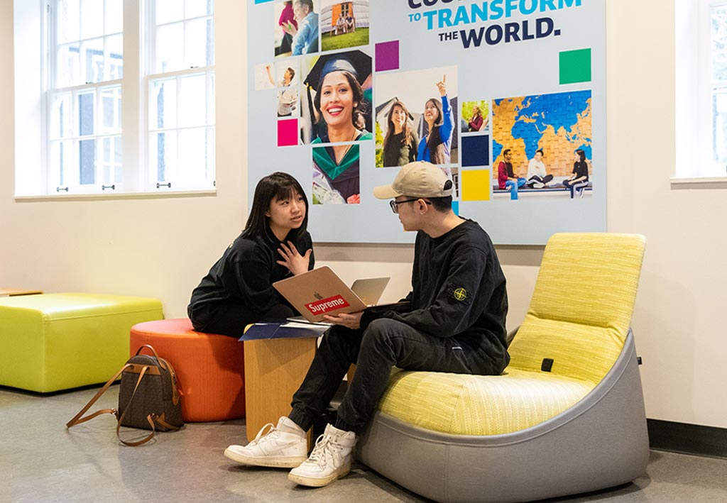 Two Royal Roads students have a conversation while sitting on colourful, lounge furniture. One holds their laptop open and the other listens intently.