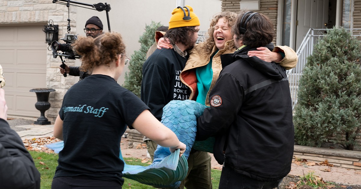 Thre people dressed in black carry a laughing woman dressed as a mermaid across a lawn.