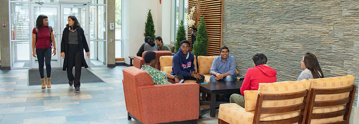 A bustling entranceway of a classroom building at Royal Roads University with students sitting around a table in conversation, and two others walking in through the front doors.