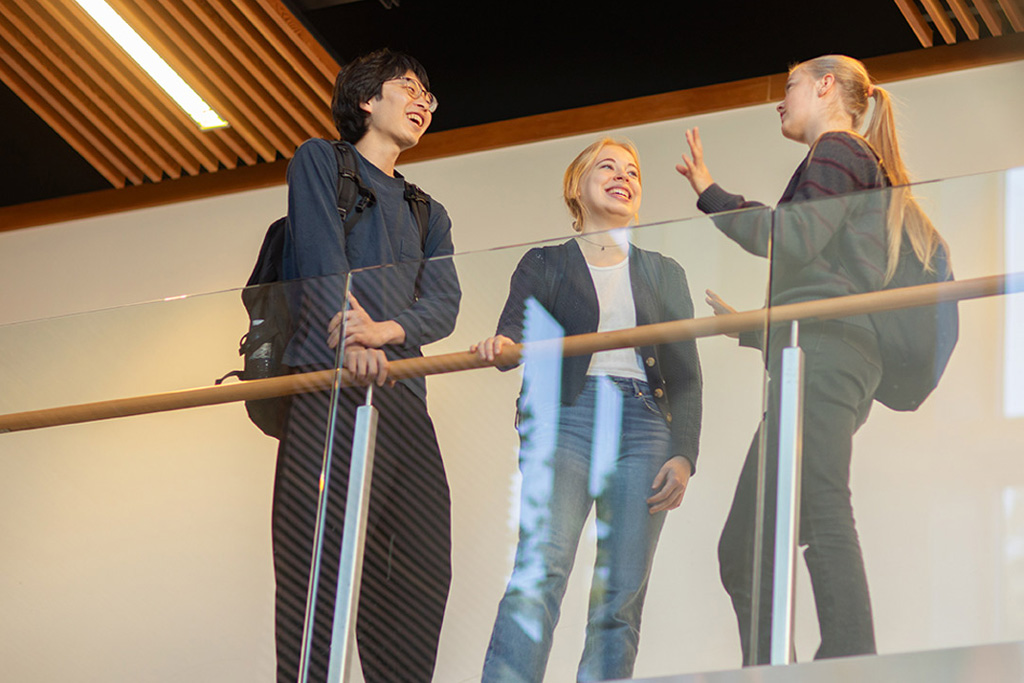 A group of students stand against a railing on the second floor of a campus building while in conversation