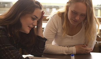 Two students studying at a desk lean forward to read a textbook side-by-side.