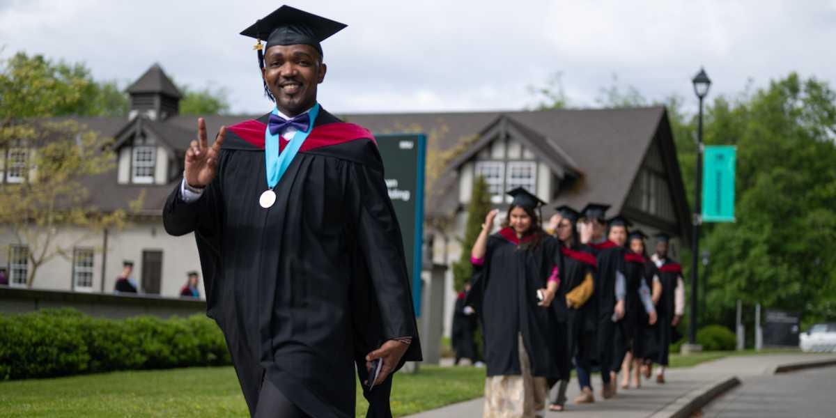 An RRU student in their graduate regalia gives the peace sign as they walk in a line with their students during a Convocation ceremony outdoors