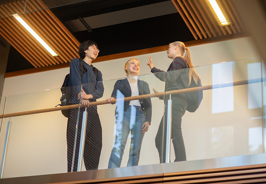 Three students gather next to a glass balcony at Royal Roads University, smiling in conversation.