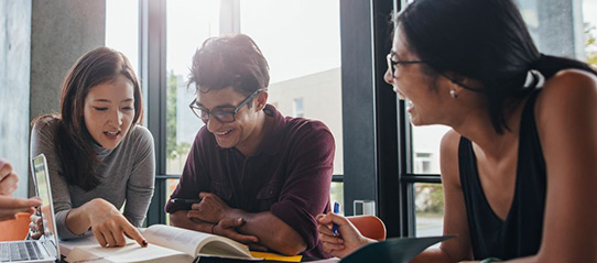 A group of four students sit at a large des, smiling as they look toward a text book.