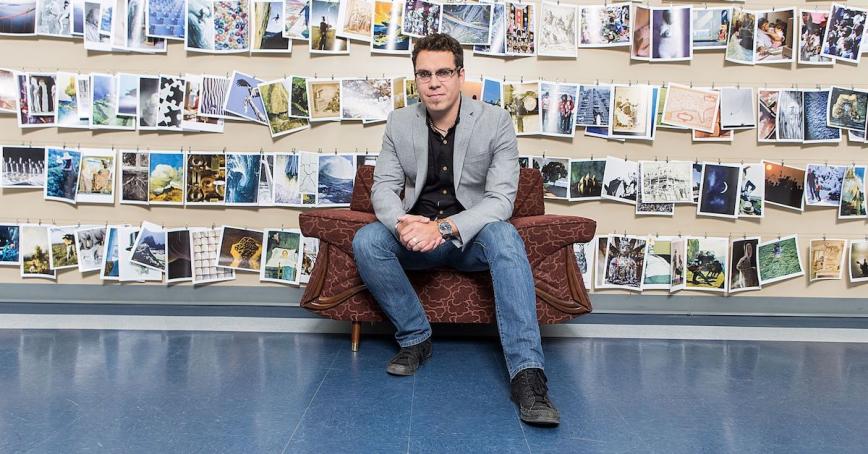 MA in Leadership graduate sits in a chair in front a wall covered in photographs.