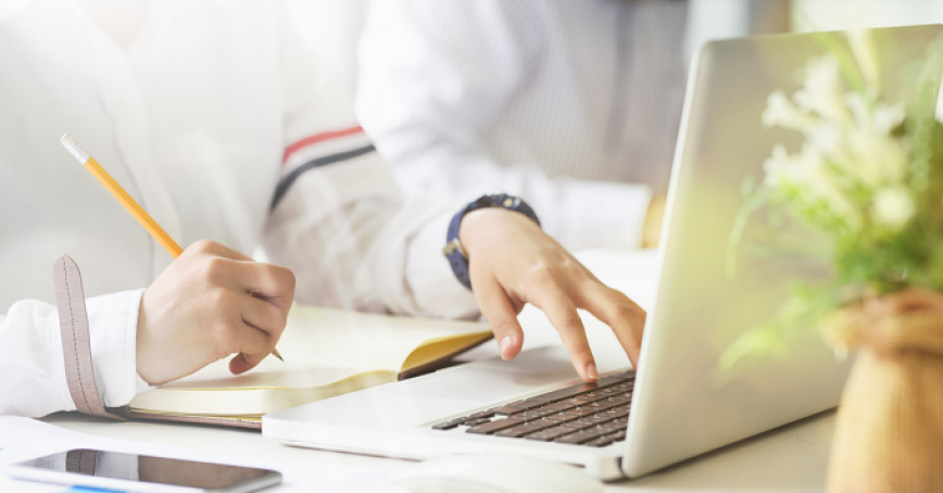 A person writing in a notebook while using a laptop