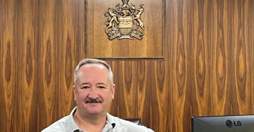 A smiling man with a moustache stands below a crest hung on a wood panel wall.