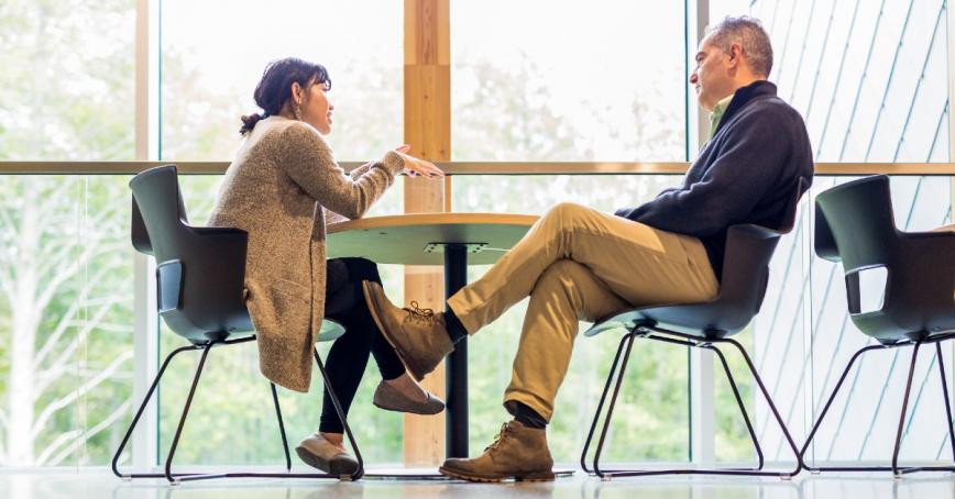 Student speaking with their instructor at a table next to a row of windows