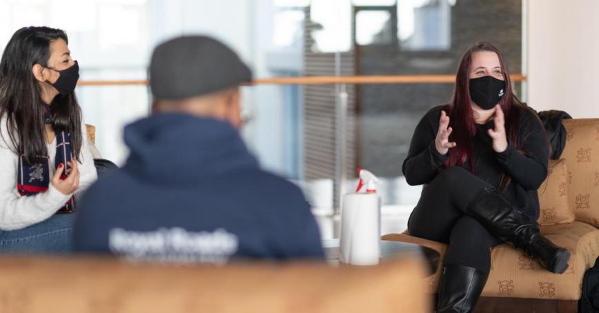 Three students sit in an open lobby. They are physically distanced and wearing masks, but quite engaged in what one students is saying.