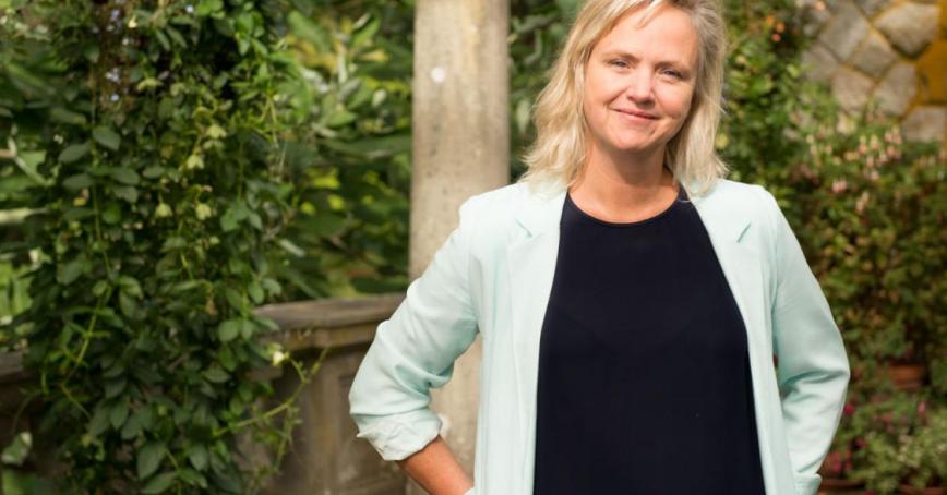 A smiling woman with blonde, shoulder-length hair wearing a mint green blazer and black t-shirt standing in front of a background of green leaves and stone columns.