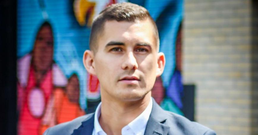 Tyson Atleo stands resolutely in front of a bright mural in a blue suit with a white open collared shirt..