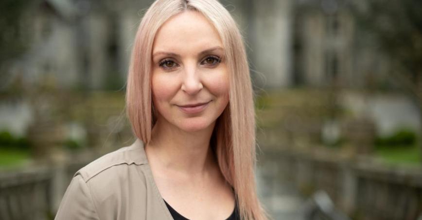 A woman with a small smile looks at the camera. A stone building is in the background.