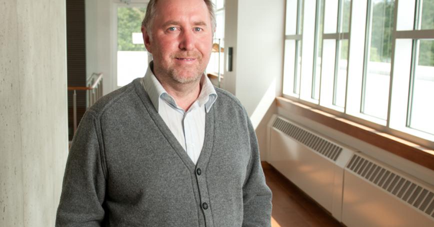 Prof. Ken Christie, standing next to a row of windows, wearing a white dress shirt and grey button up sweater