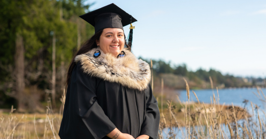 A woman stands in front of a lagoon wearing a graduation cap and gown.