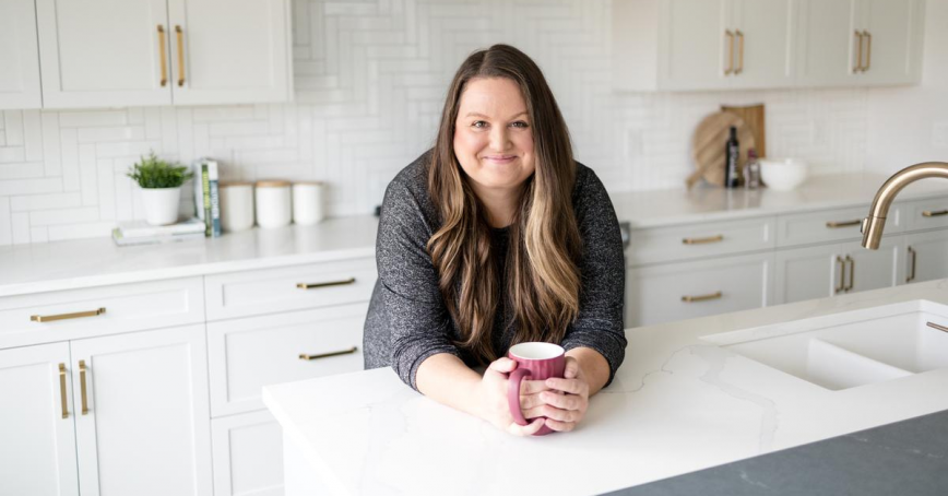 A woman rests her forearms on a kitchen island. She's cradling a coffee mug.