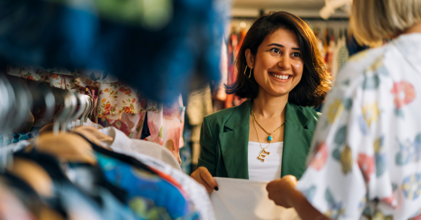 Two women smile at each other while shopping among clothes in a thrift store.