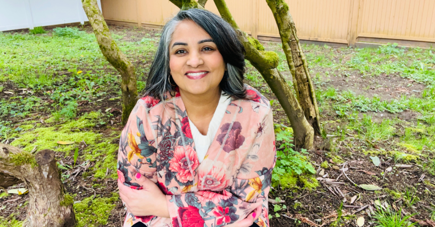 Mina Sahota sits in a garden setting, smiling at the camera.