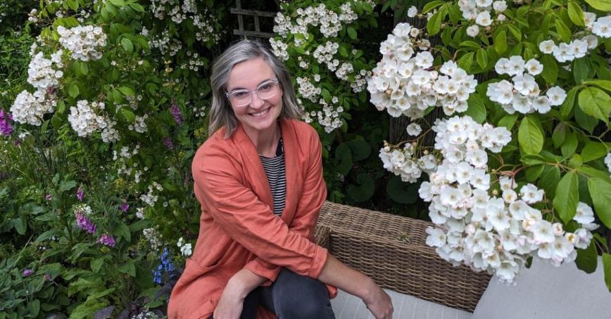 A woman wearing a coral blazer smiles out directly at the camera. She is surrounded by a blooming wisteria