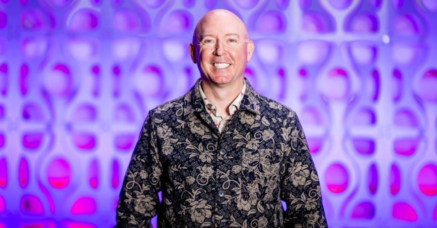 A smiling man wearing a gold patterned onyx jacket smiles out directly at the camera. He is in front of a blurred but vivid purple abstract background.