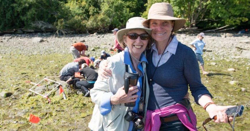 Professors Leslie King (left) and Audrey Dallimore smiling on a beach with students and partners behind doing research in clam garden.