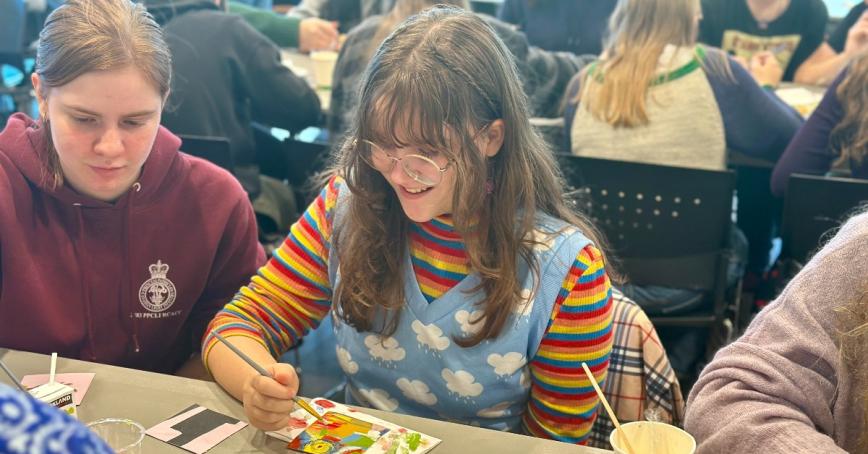 Girl painting a picture on a table, a second girl sitting next to her.