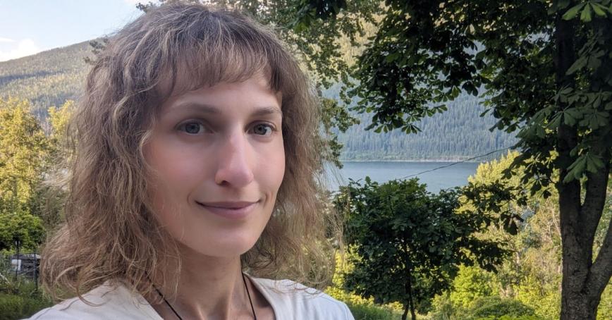 Margot Webster stands close to the camera; framed behind her is a beautiful nature scene with abundant green trees and the glistening blue of a lake in the background