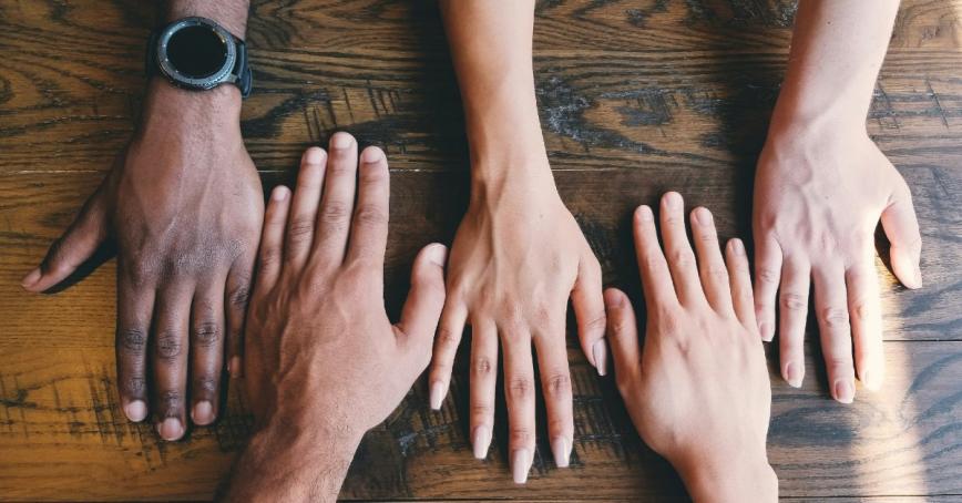 Aerial image of five hands in different skin tones laying palm down on a wooden surface.