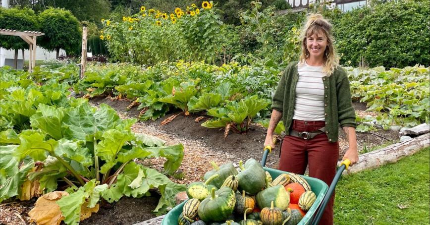 Solara Goldwynn stands in front of leafy greens at the Farm holding a wheelbarrow of squash