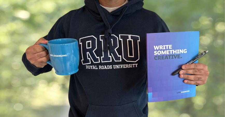 A photo of a man from the neck down holds a notebook in one hand and a mug in the other.