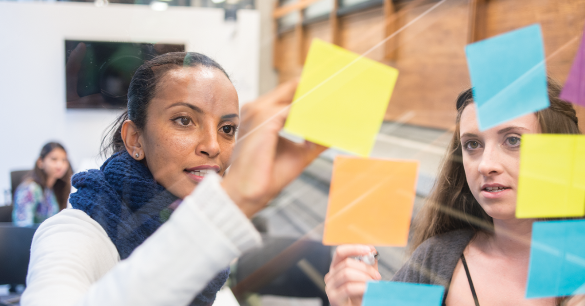 Individuals writing on and organizing colourful sticky notes on clear display board.