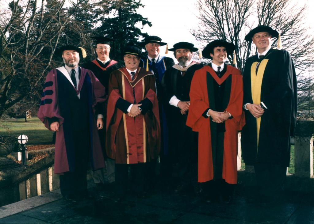 Men in academic regalia gather for a photo on a terrace outdoors. 