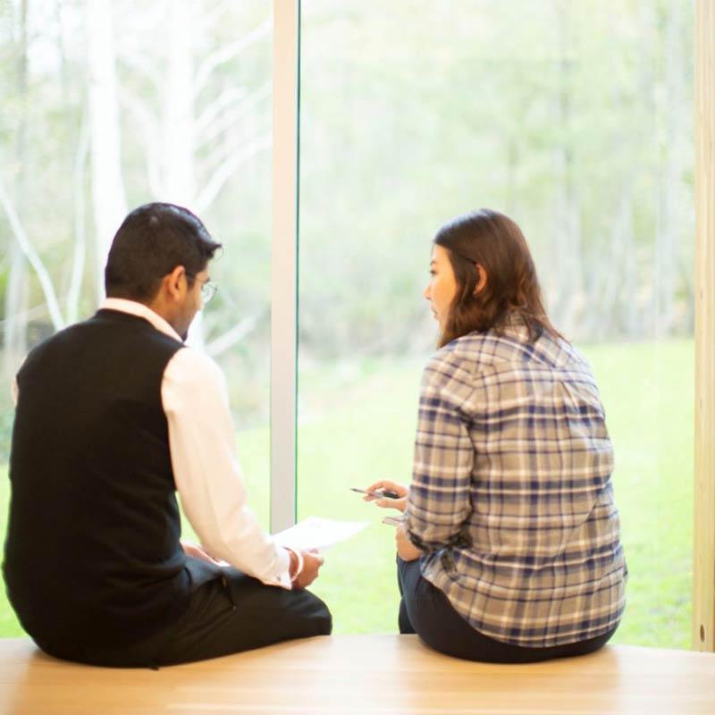 Two students sit in discussion while looking out a floor-to-ceiling window.