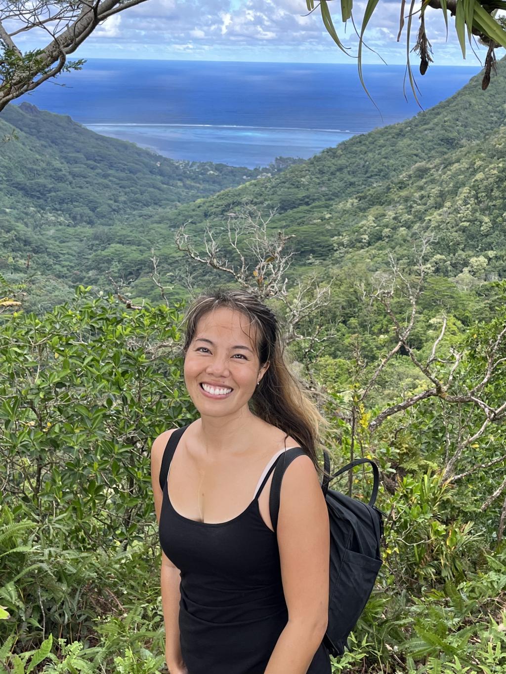 Vivian Giang stands in front of a lush green valley, with the ocean showing behind.