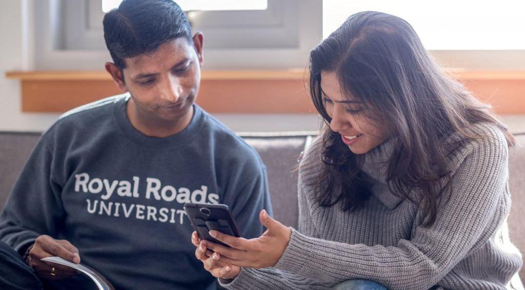 Two people sit on a couch looking at a phone while studying together.