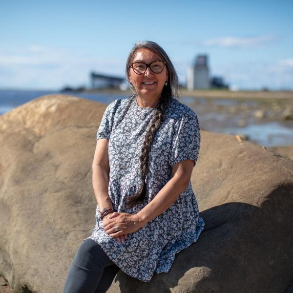 A woman sits on a large rock, smiling straight out at the camera on what looks to be a lovely summer day. A few buildings can be seen in the background.