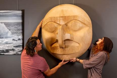 Two people lifting a carving and placing it on a gallery wall