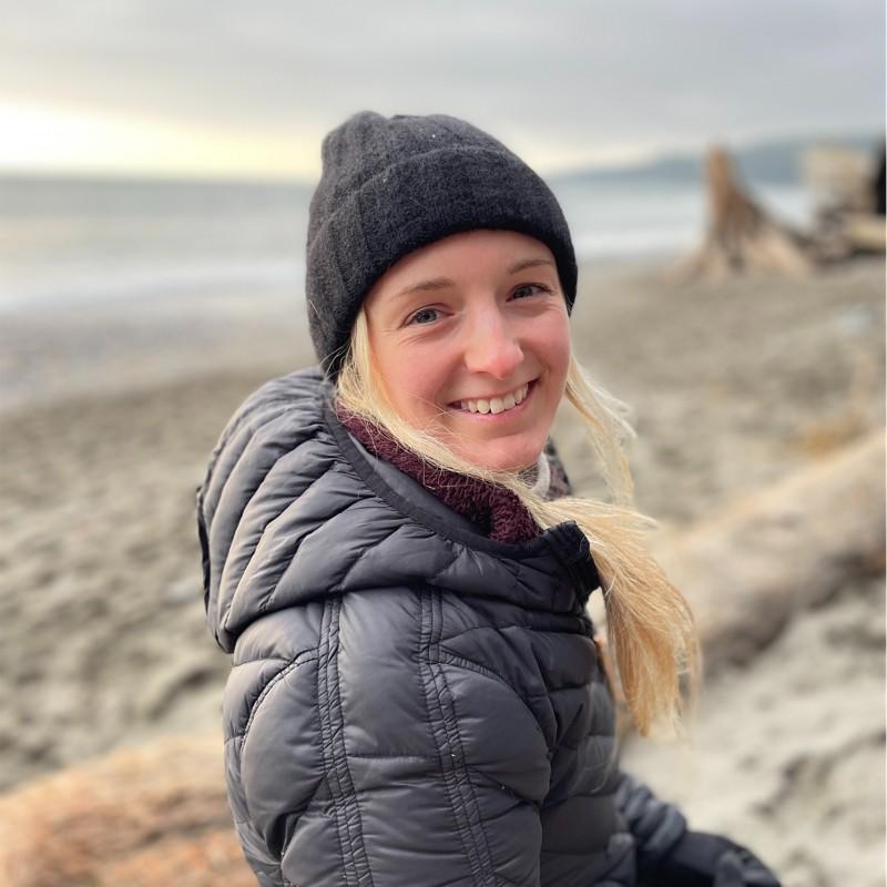 A smiling woman, wearing a black toque and jacket, looks over her shoulder at the beach.