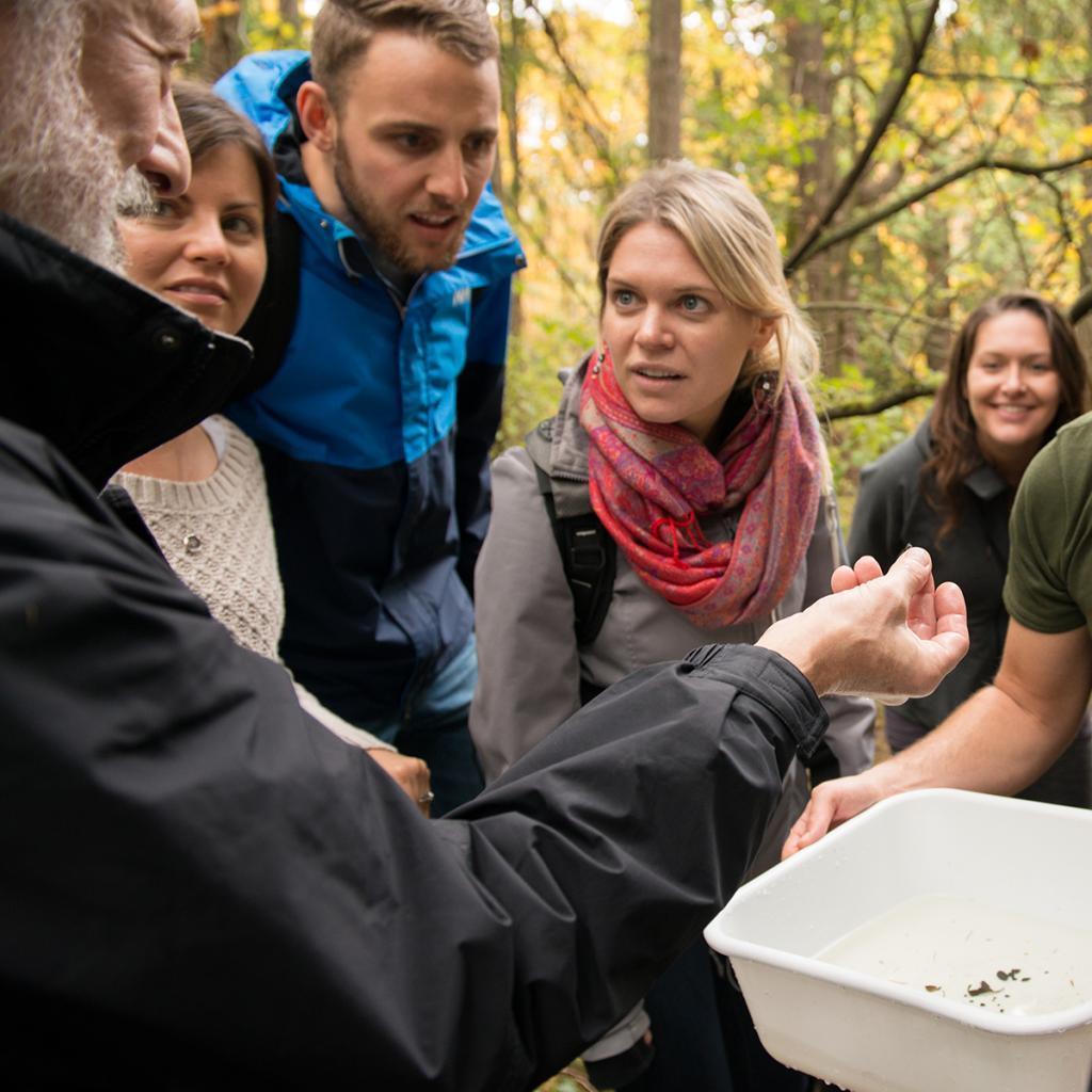 A group of master's students stand around their instructor and learn in an outdoor setting