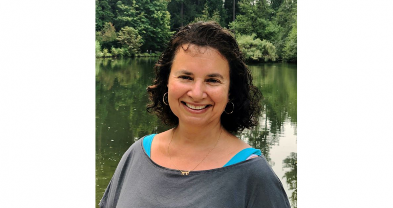 A smiling woman with shoulder-length dark hair wearing a grey sweatshirt, in front of a lake with trees in the background.