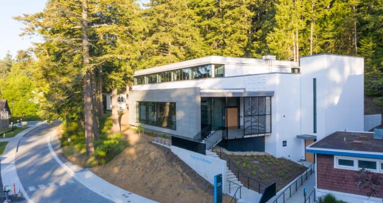 A white building with large windows is set into a forested hill on a campus.